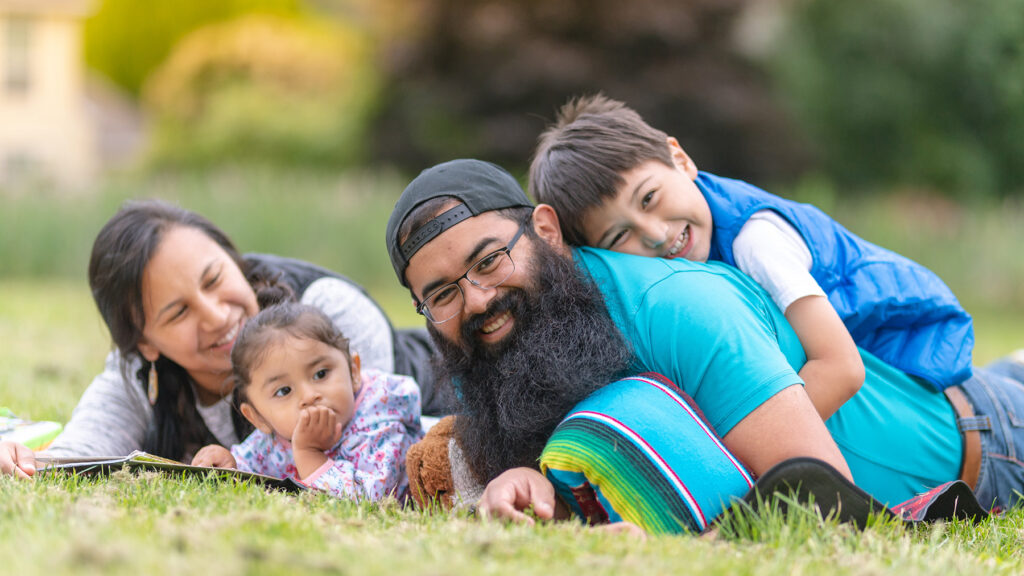 A Native American mom and dad hang out at the park and read books with their two young children on a warm summer afternoon. The boy is lying on top of his dad and they're both smiling at the camera.