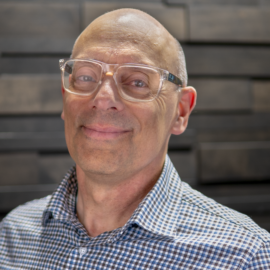 Matt Bartolotti wearing a checkered blue, black, and white button up shirt. He is standing in front of a dark grey background.